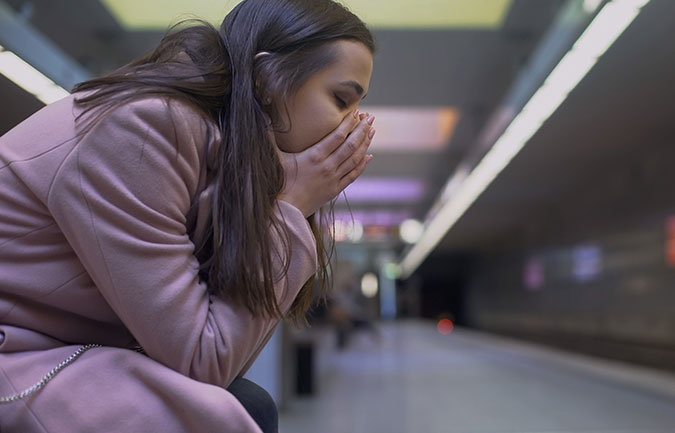 woman in pink coat sat at train station platform with head in hands