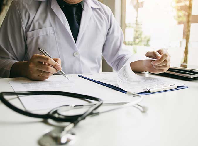Photo of a person sitting at a desk, with a pen in their right hand and a clipboard of paperwork in their left hand. They are wearing a white clinician's coat. A stethoscope lies on the desk.