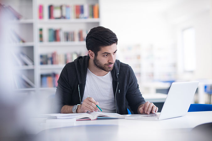 man in a library workspace using laptop and taking notes