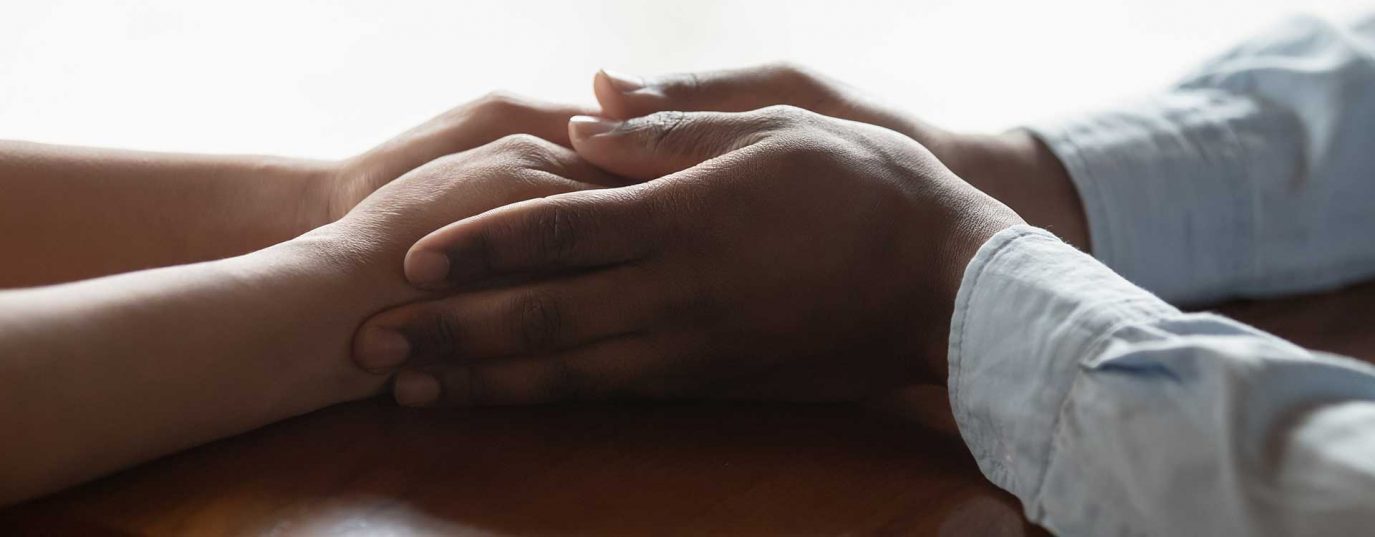 two people with hands on the table, one supportively holding the others